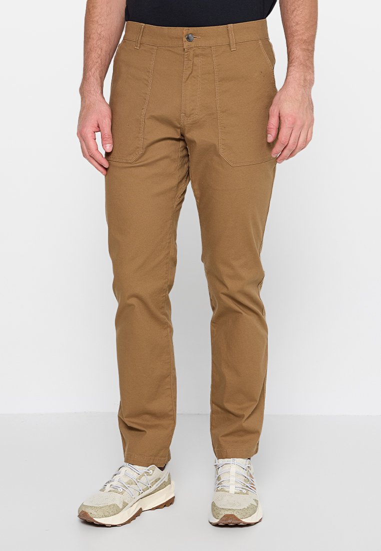 Hombre vestido con pantalones rectos marrones, con bolsillos frontales y zapatillas beigas, de pie frente a un fondo blanco liso.