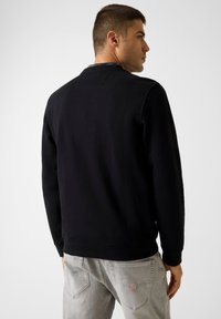 Fekete pulóver kerek nyakkal, puha anyagból készült. Bordázott mandzsettákkal és szegéllyel rendelkezik. Szürke farmernadrág hátulján logó található.