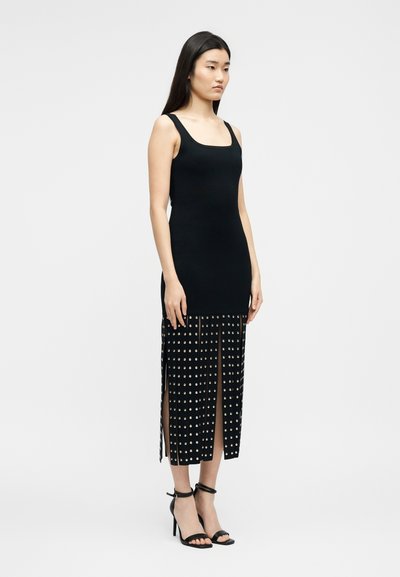 Γυναίκα με μαύρο φόρεμα χωρίς μανίκια, με φούστα με σκίσιμο και διακοσμητικά καρφιά, φορώντας μαύρες ψηλοτάκουνες σανδάλες, στέκεται μπροστά σε μονόχρωμο φόντο.