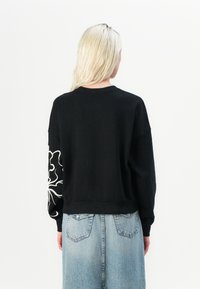 Felpa nera con vestibilità rilassata, caratterizzata da un design floreale bianco sulla manica sinistra. Abbinata a pantaloni in denim blu chiaro.