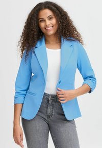 Světle modrý blazer z hladkého materiálu, s jednokneprovým zapínáním, klopovými límci a vyhrnutými rukávy, v kombinaci s bílým topem a šedými džíny.