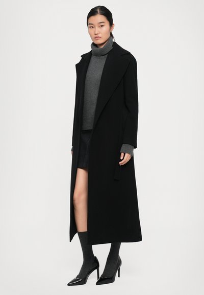 Woman wearing a long black coat, gray turtleneck sweater, gray knee-high socks, black high heels, and a black skirt standing against a white background.