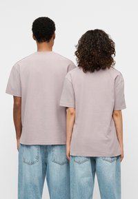 Δύο άτομα φορούν μεγάλα, λιλά μπλουζάκια από μαλακό βαμβάκι, συνδυασμένα με ανοιχτόχρωμα, φαρδιά τζιν παντελόνια. Απλός σχεδιασμός, χωρίς ορατά λογότυπα.