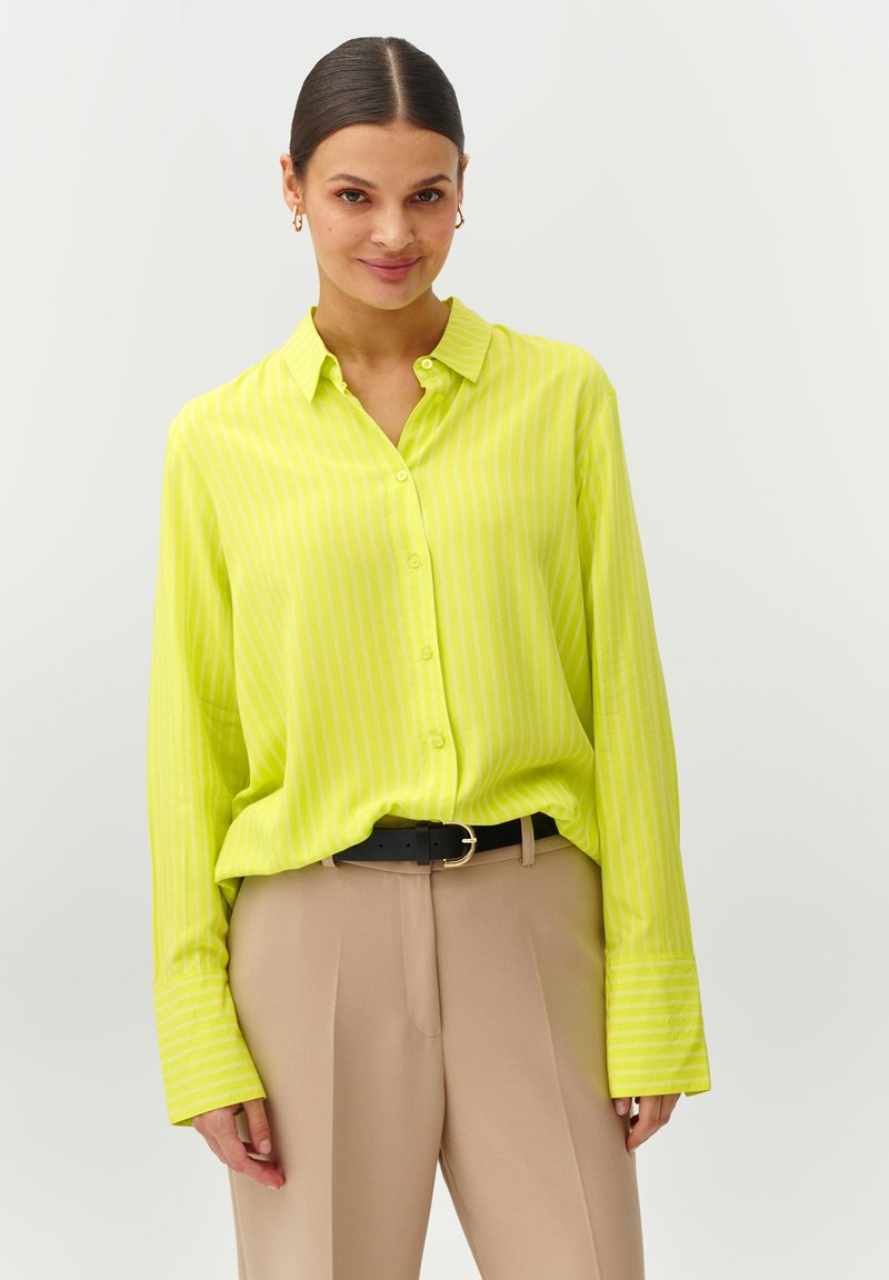 Camisa de botón de un brillante verde lima con rayas verticales blancas, con cuello y mangas largas, combinada con pantalones beiges y un cinturón negro.