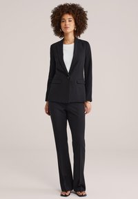 Kobieta o kręconych włosach, ubrana w czarny, dopasowany garnitur z białą bluzką i czarnymi sandałami, stojąca na jednolitym tle.