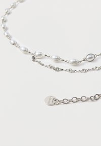 Colier stratificat cu lanț din argint, perle albe și accente de cristal. Include o etichetă rotundă cu numele brandului lângă închizătoare.