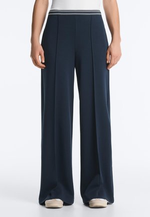 Femeie purtând pantaloni largi bleumarin cu talie elastică și adidași albi, stând în picioare pe un fundal simplu.