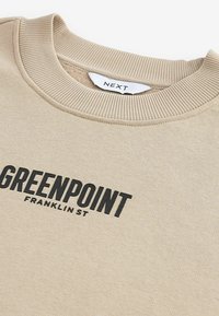 Bež dukserica s rebrastim ovratnikom, na prednjoj strani ima veliki crni natpis "GREENPOINT FRANKLIN ST". Glatka tekstura.