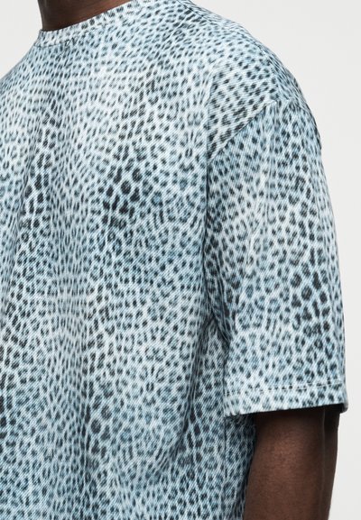 Άνδρας που φοράει κοντομάνικο πουκάμισο με ακανόνιστο μοτίβο σε ανοιχτό μπλε και μαύρο, φωτογραφημένος από τους ώμους έως το μέσο του κορμού.