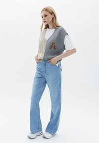 Gilet lavorato a maglia in grigio e crema con motivi a trecce e un dettaglio con la lettera "A", indossato sopra una t-shirt bianca, abbinato a jeans azzurri chiari.