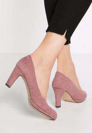 Feet wearing mauve suede high-heeled shoes paired with black cropped pants with side slits on a white background.