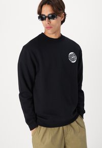 Zwarte sweatshirt met geribde manchetten en onderkant, voorzien van een circulaire witte graphic op de linkerborst. Model draagt een donkere zonnebril.