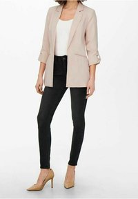 Beige skräddarsydd blazer med uppvikta ärmar, kombinerad med svarta skinny jeans och nakna spetsiga klackar. Släta texturer och åtsittande design.