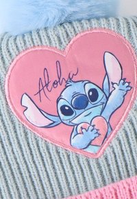 Grijze gebreide beanie met een roze hartpatch waarop een blauwe figuur met een hart staat, afgewerkt met lichtblauwe nepbont aan de bovenkant.