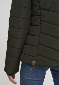Dark green puffer jacket with horizontal quilted texture, brown label detail at the hem, and ribbed cuffs, paired with blue denim jeans.