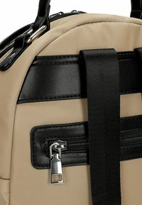 Sac à dos en tissu beige avec des accents en cuir synthétique noir, équipé d'une poche zippée à l'avant et de bretelles noires réglables. Détails en métal argenté.