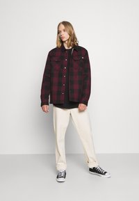 Chemise en flanelle à carreaux bordeaux et noirs avec deux poches poitrine, portée sur un t-shirt noir, assortie à un pantalon crème et des baskets noires.