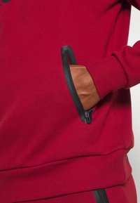 Sweatshirt rouge en tissu doux avec une poche zippée noire sur le côté et des poignets côtelés, offrant un ajustement confortable.