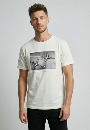 Homme aux cheveux courts portant un t-shirt blanc avec un graphisme noir et blanc de deux avions à réaction en plein vol sur un fond gris.