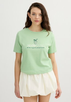 Joven mujer con cabello largo y castaño lleva una camiseta verde claro con la inscripción "Slow Living" y una falda blanca, de pie contra un fondo claro y liso.