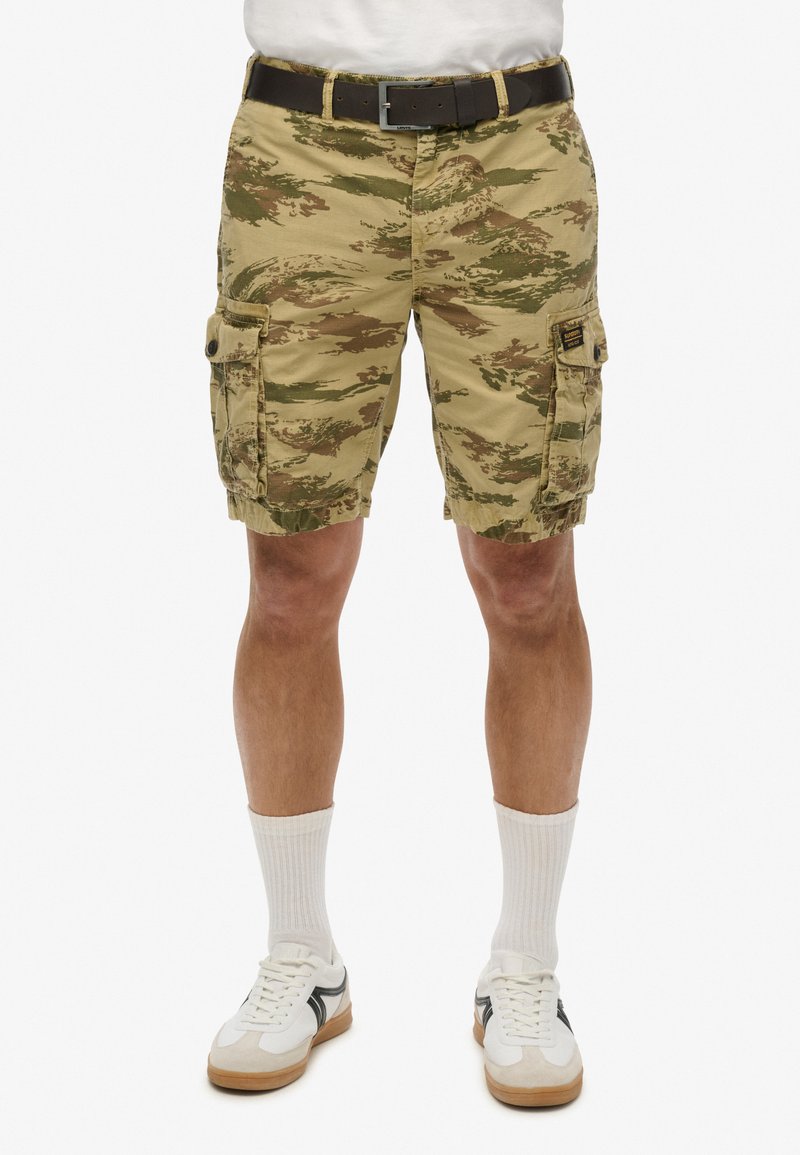 Camo cargo shorts i khaki, grøn og brun med lommer, et bælte og en afslappet pasform. Båret med hvide sokker og sneakers.