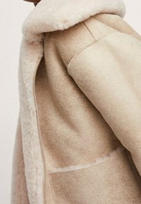 Abrigo de borrego beige con un suave cuello de lana visible y forro interior, mostrando una textura detallada y una cremallera a lo largo del borde frontal.
