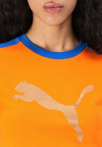 Pomarańczowy bawełnowy t-shirt z niebieskim prążkowanym dekoltem, z logo Puma ozdobionym srebrnymi kryształkami. Gładka tekstura, luźny krój z krótkimi rękawami.