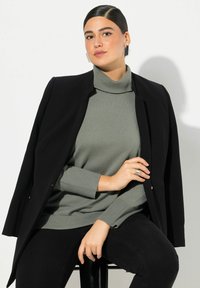 Schwarze Blazer über einem grauen Rollkragenpullover mit gerippten Bündchen. Das Modell sitzt auf einem schwarzen Hocker mit einem zarten Ring am Handgelenk und minimalistischen Ohrringen.