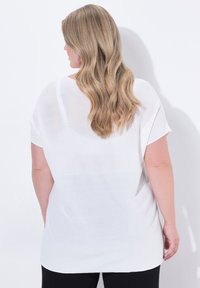 Top bianco in maglia trasparente con maniche corte e vestibilità rilassata, caratterizzato da un orlo arrotondato e una texture sottile, abbinato a pantaloni neri.