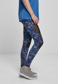 Leggings imaju digitalni kamuflažni uzorak u plavoj, ljubičastoj i crnoj boji s mrežastim panelima. Upareni su sa sivim visoko-topericama.