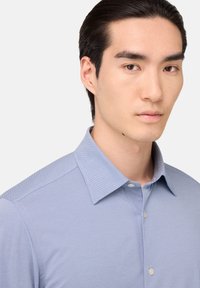 Camicia azzurra chiara con un sottile motivo a righe, colletto aperto e una texture liscia. Presenta una chiusura con un solo bottone al collo.