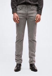 Άνδρας που φοράει ξεθωριασμένο γκρι στενό τζιν, μαύρα μποτάκια αστραγάλου και σκούρο τζιν πουκάμισο με τα χέρια χαλαρά στα πλάγια.