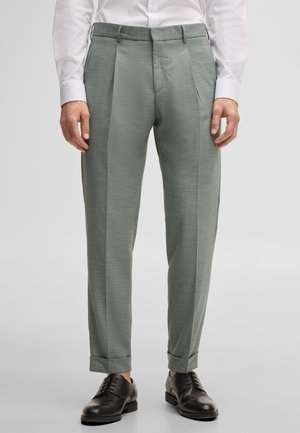 Man draagt lichtgrijze op maat gemaakte broek met omslagen, een wit overhemd en zwarte leren nette schoenen, staand tegen een effen achtergrond.