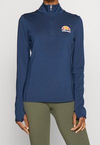 Marinblå långärmad topp med kvartslång dragkedja, mjukt tyg, med en broderad flerfärgad logotyp på bröstet och tumhål vid ärmsluten.