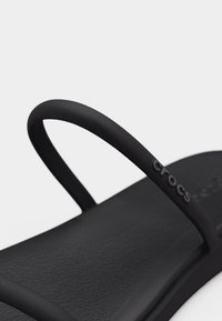 Μαύρο σανδάλι Crocs με λεπτά λουριά και υφή στην επιφάνεια του πέλματος, που φέρει το λογότυπο Crocs στο πλευρικό λουράκι.