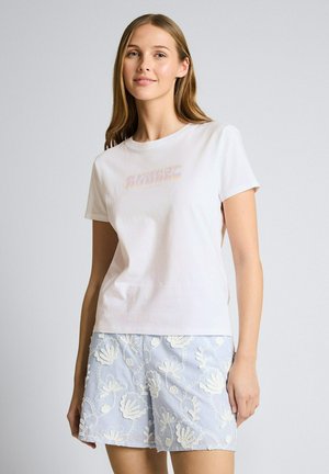 Vrouw met lang haar, gekleed in een wit T-shirt met korte mouwen en lichtblauwe shorts met witte bloemembroidery, staand tegen een effen achtergrond.