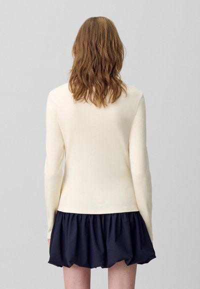 Personne avec des cheveux bruns ondulés de dos, portant un haut à manches longues crème et une jupe courte bleu marine froncée sur un fond uni.