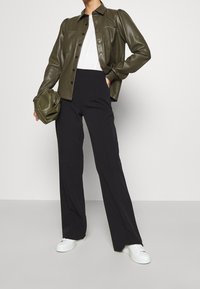 Giacca camicia in ecopelle verde oliva con spalle a palloncino e quattro tasche frontali, abbinata a pantaloni neri a gamba larga e sneakers bianche.