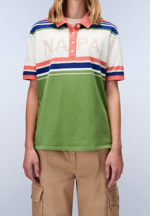 Person iført en kortærmet polo med grønne, hvide, blå og koralfarvede vandrette striber og beige cargobukser foran en ensfarvet baggrund.