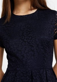 Námořnické modré krajkové šaty se složitým květinovým vzorem, krátkými rukávy a upnutým korzetem, který přechází do skládánové sukně.