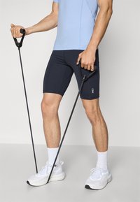 Kék, testhezálló sportfelső hálós díszítésekkel, sötétkék rövidnadrág és fehér sportcipő. A felhasználó fekete fitness ellenállásgumikat tart, markolattal.