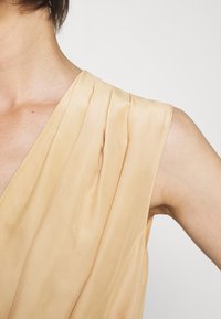 Femme portant un chemisier sans manches en satin beige avec des détails plissés près de l'épaule, sur un fond blanc uni.