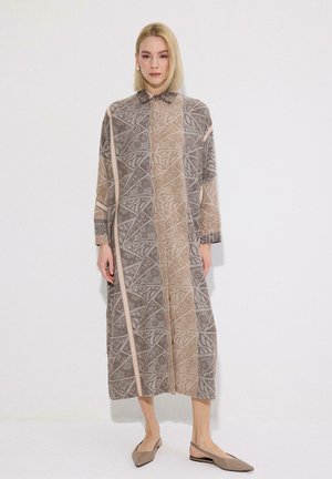 Žena so svetlými vlasmi, ktorá nosí dlhé šaty so vzorom geometrických tvarov v béžovej a sivej farbe, v kombinácii s béžovými balerínami, stojí pri bielej stene.