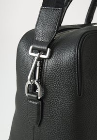 Geantă din piele neagră cu un finisaj texturat, având un fermoar și accesorii din metal argintiu, precum și o curea lată ajustabilă pentru flexibilitate.