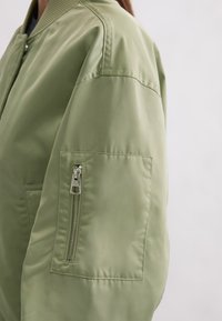 Light green bomber jacket with a ribbed collar and a zippered pocket on the left sleeve. Smooth, glossy fabric with a relaxed fit.