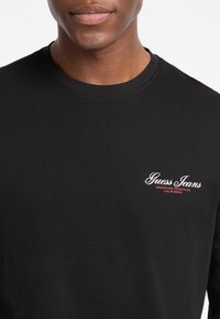 Sort langærmet T-shirt med rund halsudskæring og et lille hvidt og rødt "Guess Jeans" logo på brystet. Blød bomuldsstruktur.