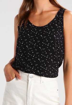 Woman wearing a black sleeveless top with small white star patterns and high-waisted white pants, hand in pocket.