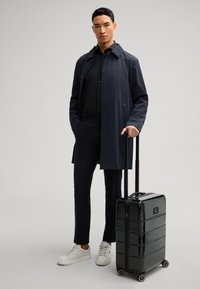 Valigia rigida nera con superficie liscia, scanalature verticali e quattro ruote. L'utente indossa un lungo cappotto scuro, una maglia scura e sneakers bianche.