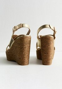 Sandales compensées dorées à lanières avec une plateforme et un talon en liège. Les caractéristiques incluent une sangle à boucle et une finition métallique lisse.