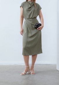 Olivengroene jurk met een gedrapeerde halslijn, een getailleerde taille en midi-lengte. Gecombineerd met beige sandalen met bandjes en een zwarte clutch. Soepele textuur.